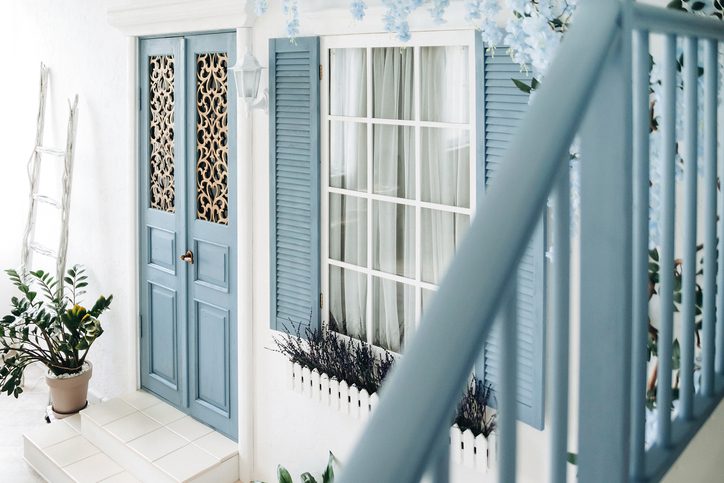 Blue door and window on the white wall of a mediterranean-style house with a flowering tree weaving along the wall and potted plants. Porch of a house in Santorini